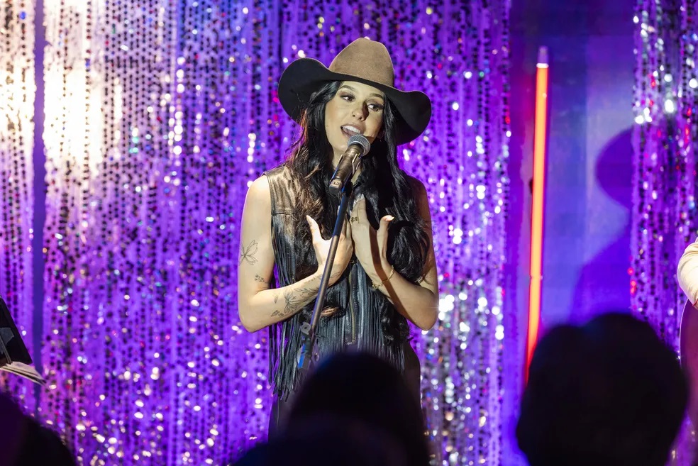 Ana Castela cantando em microfone, de roupa marrom e chapéu, de cabelo solto