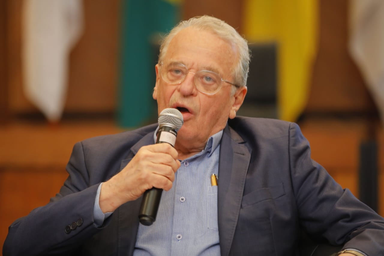 Tarso Genro falando em microfone, de roupa social, sério, de óculos de grau, sem olhar para a câmera