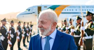 O presidente Luiz Inácio Lula da Silva (PT) sorrindo e olhando para o lado, com avião ao fundo