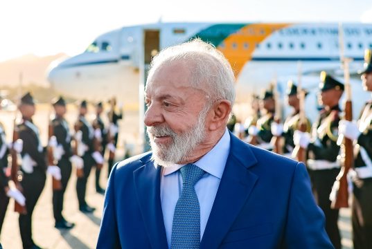 O presidente Luiz Inácio Lula da Silva (PT) sorrindo e olhando para o lado, com avião ao fundo
