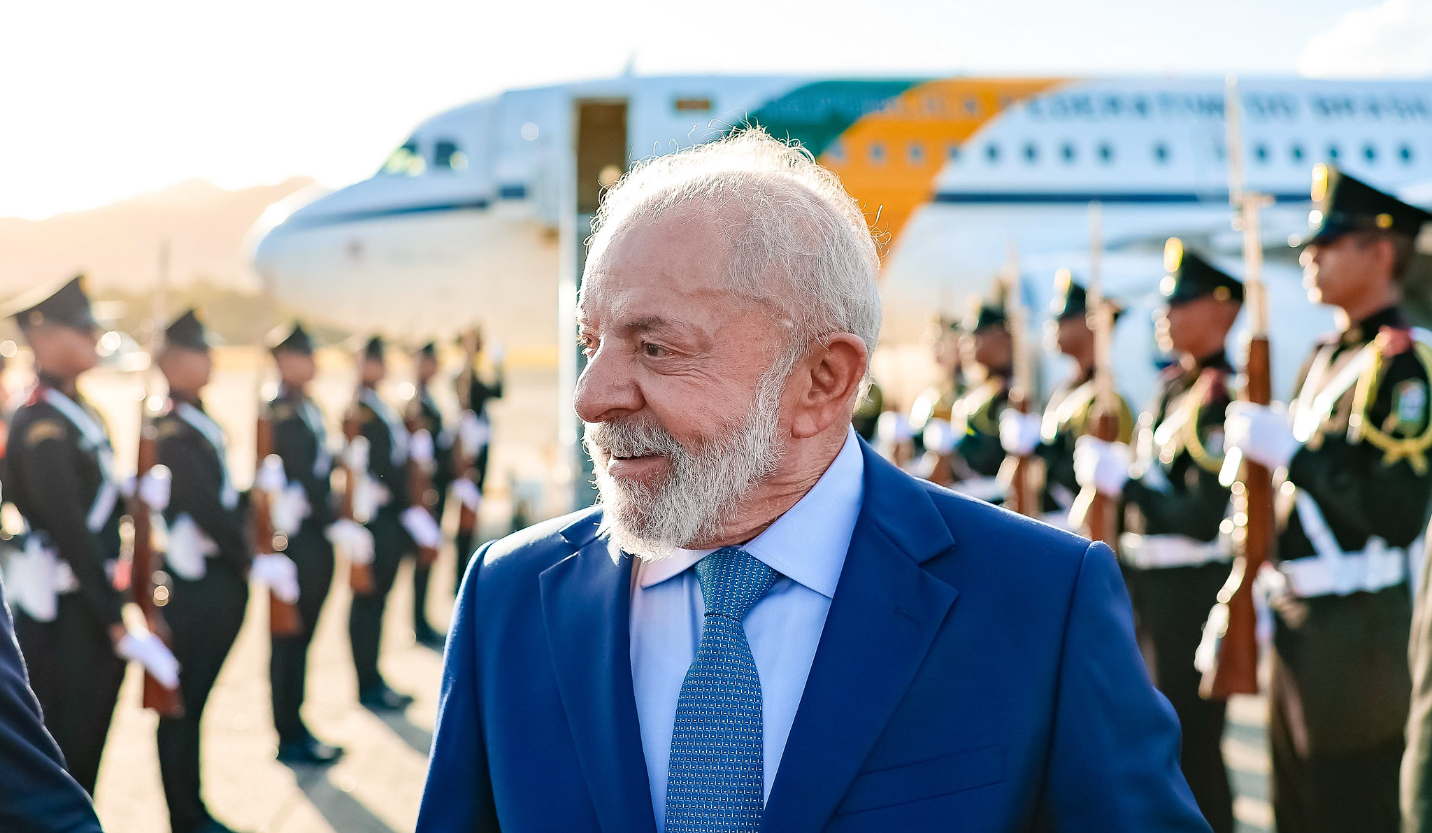 O presidente Luiz Inácio Lula da Silva (PT) sorrindo e olhando para o lado, com avião ao fundo