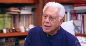 Antonio Fagundes de roupa de frio azul marinho, falando, sério, sem olhar para a câmera