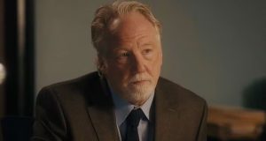 O ator e diretor Timothy Busfield sério, sem olhar para a câmera, de roupa social marrom