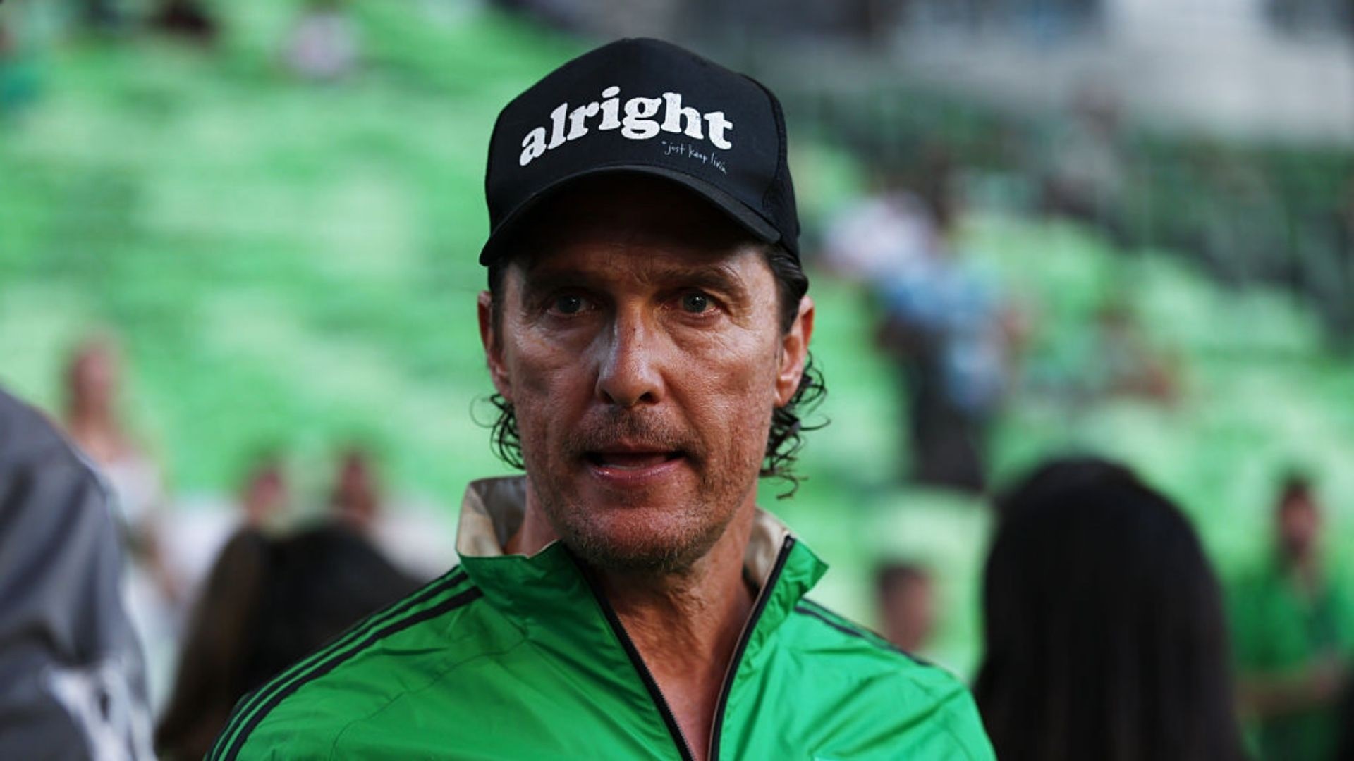O ator americano Matthew McConaughey sério, de boné preto e roupa verde