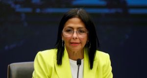 Delcy Rodríguez, presidente interina da Venezuela, de óculos de grau e roupa social branca e amarela, falando em microfone, de cabelo solto, sem olhar para a câmera