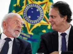 Lula e o ministro da Educação, Camilo Santana, conversando, sentados lado a lado, com bandeiras ao fundo