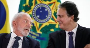 Lula e o ministro da Educação, Camilo Santana, conversando, sentados lado a lado, com bandeiras ao fundo