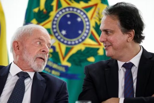Lula e o ministro da Educação, Camilo Santana, conversando, sentados lado a lado, com bandeiras ao fundo