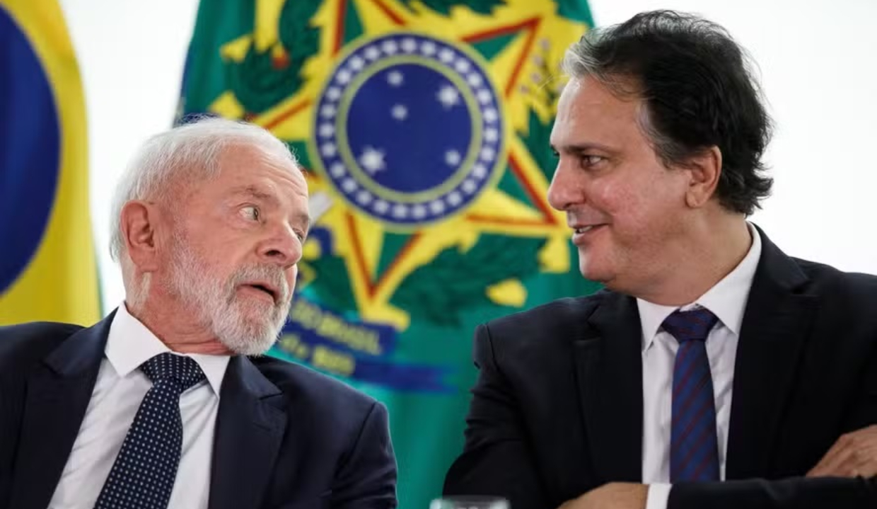 Lula e o ministro da Educação, Camilo Santana, conversando, sentados lado a lado, com bandeiras ao fundo