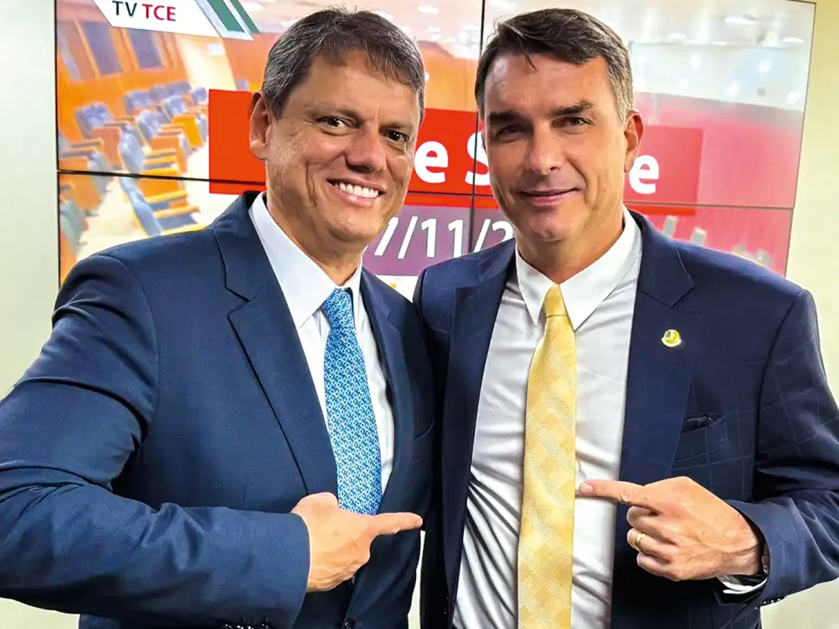 Tarcísio de Freitas e Flávio Bolsonaro posando lado a lado e apontando um para o outro