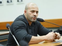 Daniel Silveira sentado, de camisa social preta, sério, falando em microfone