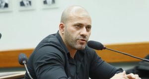 Daniel Silveira sentado, de camisa social preta, sério, falando em microfone