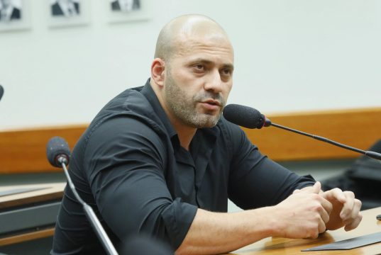 Daniel Silveira sentado, de camisa social preta, sério, falando em microfone