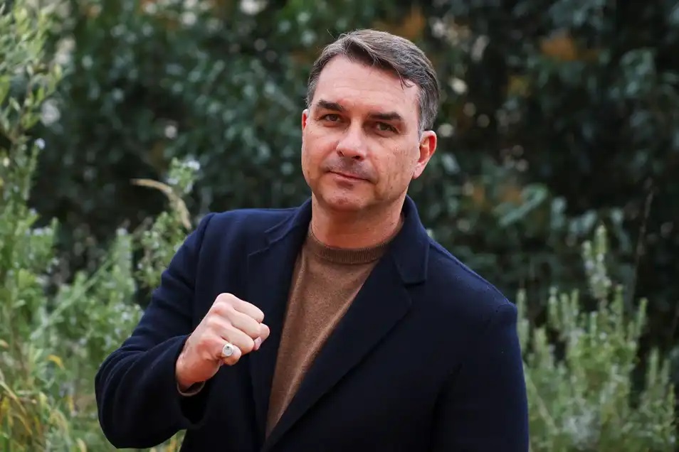 o senador Flávio Bolsonaro de roupa preta e marrom, posando para foto com as mãos fechadas