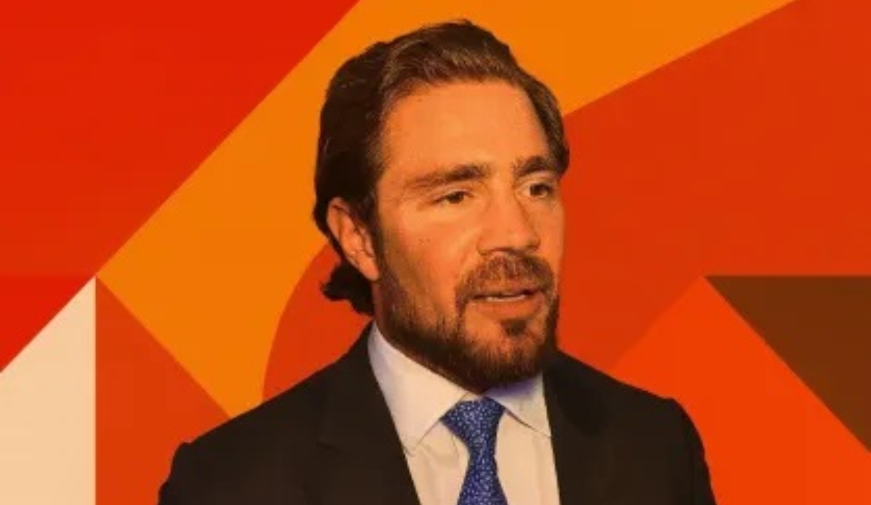 Daniel Vorcaro, controlador do Banco Master, falando, perto de painel com tons de laranja, sério, sem olhar para a câmera, de roupa social