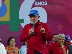 O presidente Lula (PT) de camisa vermelha e boné azul, em pé, falando em microfone