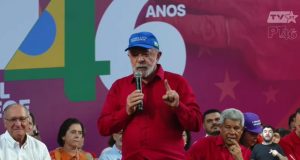 O presidente Lula (PT) de camisa vermelha e boné azul, em pé, falando em microfone