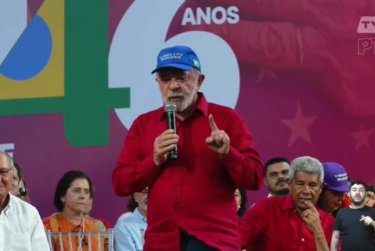 O presidente Lula (PT) de camisa vermelha e boné azul, em pé, falando em microfone
