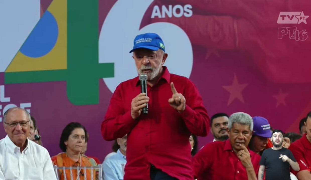 O presidente Lula (PT) de camisa vermelha e boné azul, em pé, falando em microfone