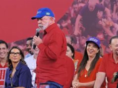 O presidente Luiz Inácio Lula da Silva (PT) de camisa social vermelha e boné azul, em pé, andando e falando em microfone, sério, sem olhar para a câmera