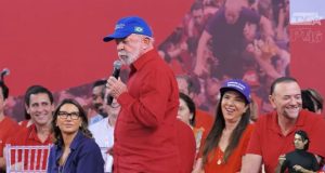 O presidente Luiz Inácio Lula da Silva (PT) de camisa social vermelha e boné azul, em pé, andando e falando em microfone, sério, sem olhar para a câmera