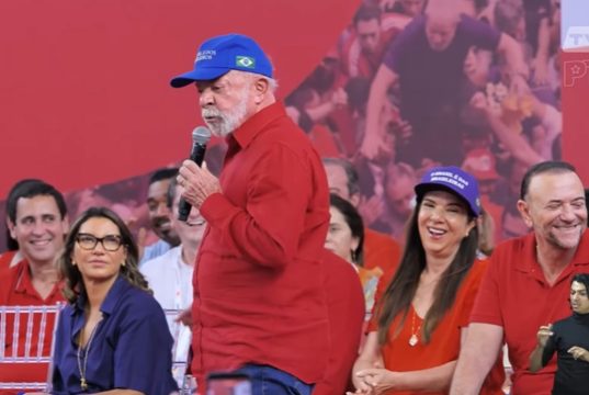 O presidente Luiz Inácio Lula da Silva (PT) de camisa social vermelha e boné azul, em pé, andando e falando em microfone, sério, sem olhar para a câmera