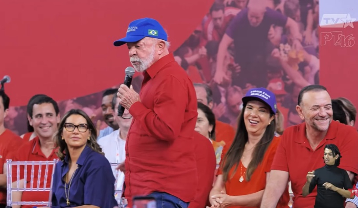 O presidente Luiz Inácio Lula da Silva (PT) de camisa social vermelha e boné azul, em pé, andando e falando em microfone, sério, sem olhar para a câmera