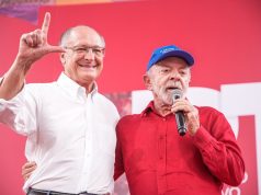O vice-presidente Geraldo Alckmin e o presidente Lula no aniversário do PT, em pé, abraçados, o primeiro sorrindo e o segundo falando em microfone