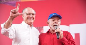 O vice-presidente Geraldo Alckmin e o presidente Lula no aniversário do PT, em pé, abraçados, o primeiro sorrindo e o segundo falando em microfone