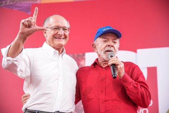 O vice-presidente Geraldo Alckmin e o presidente Lula no aniversário do PT, em pé, abraçados, o primeiro sorrindo e o segundo falando em microfone