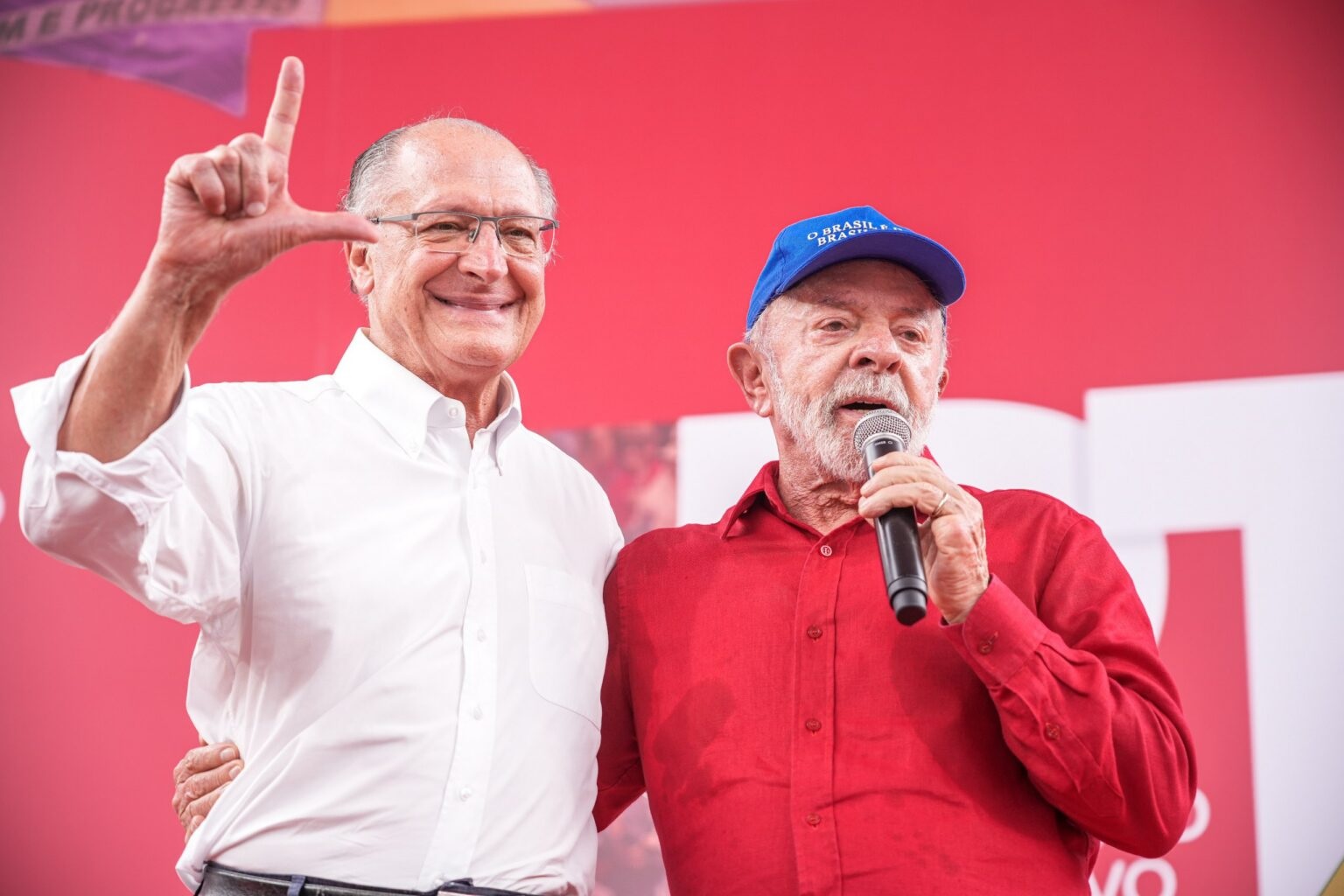 O vice-presidente Geraldo Alckmin e o presidente Lula no aniversário do PT, em pé, abraçados, o primeiro sorrindo e o segundo falando em microfone