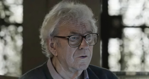 O ator Juca de Oliveira sério, de óculos de grau, em cena, sem olhar para a câmera