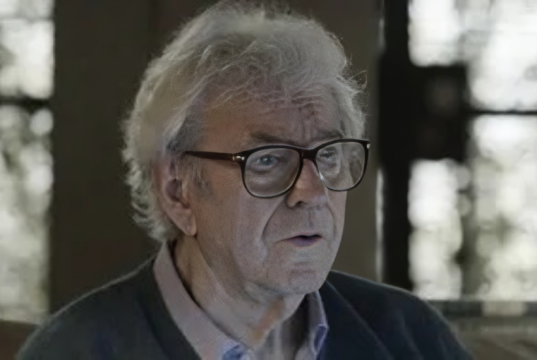 O ator Juca de Oliveira sério, de óculos de grau, em cena, sem olhar para a câmera