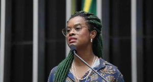 A deputada federal Carol Dartora (PT-PR) falando em microfone, séria