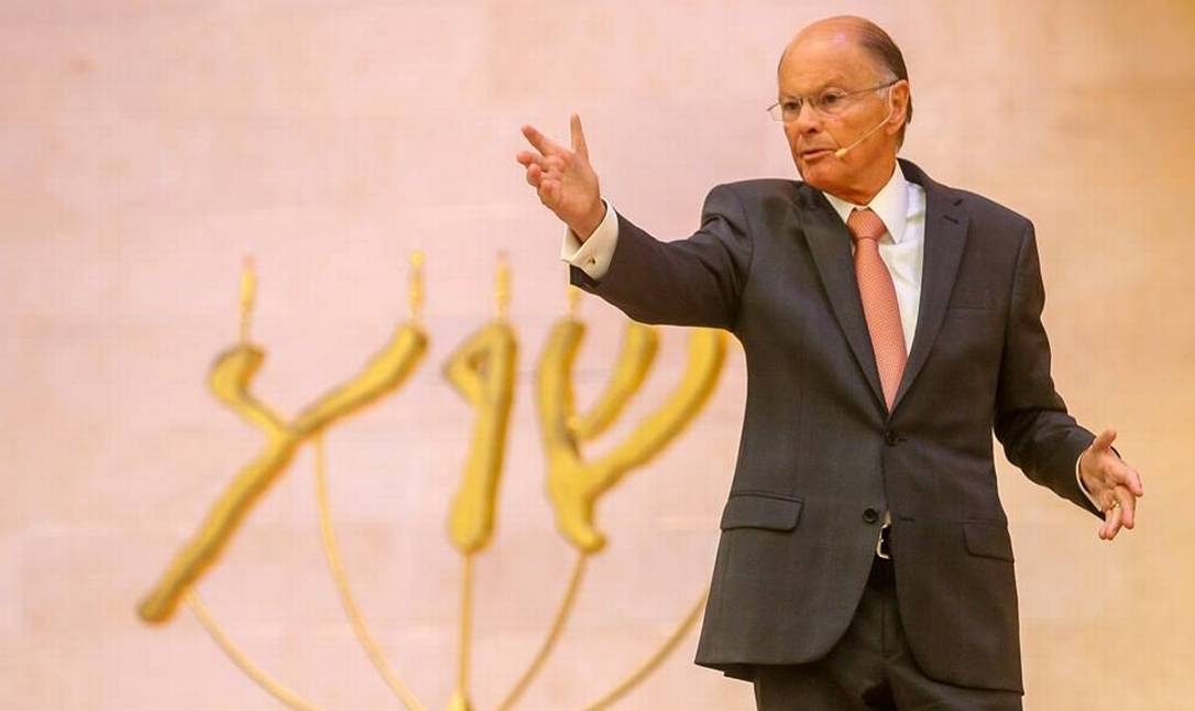 Bispo Edir Macedo falando, em pé, sério, gesticulando, sem olhar para a câmera, de roupa social e óculos de grau