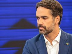 Eduardo Leite sério, olhando para o lado, no canto direito de foto