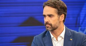 Eduardo Leite sério, olhando para o lado, no canto direito de foto