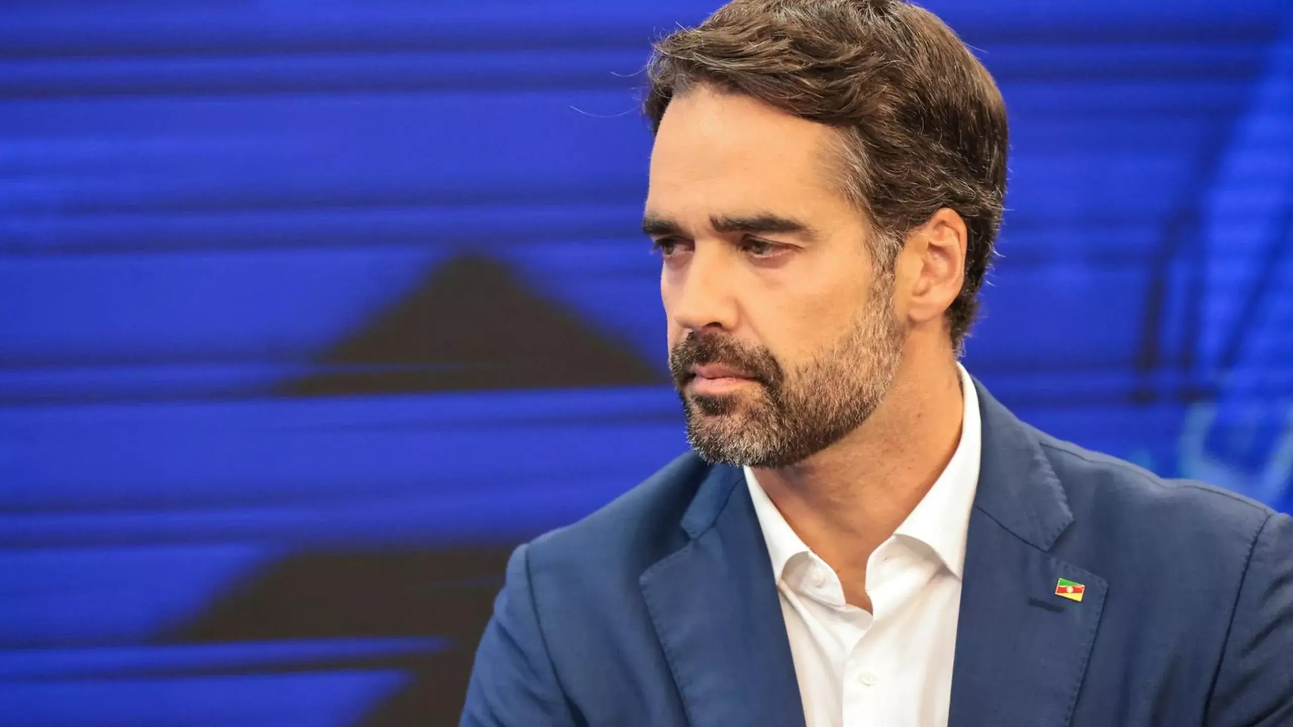 Eduardo Leite sério, olhando para o lado, no canto direito de foto