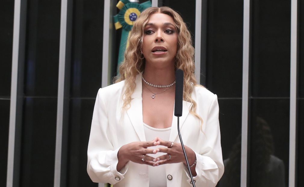 deputada federal Erika Hilton de roupa branca e cabelo solto, falando, em pé, séria