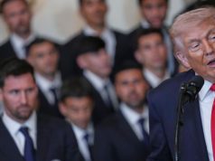 O presidente dos Estados Unidos, Donald Trump, falando, sendo observado por outras pessoas, no canto direito de foto