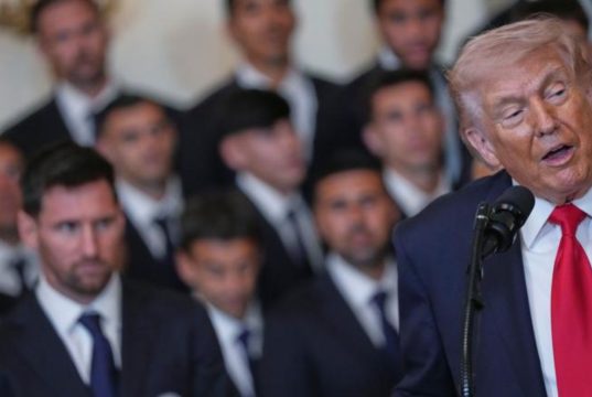 O presidente dos Estados Unidos, Donald Trump, falando, sendo observado por outras pessoas, no canto direito de foto