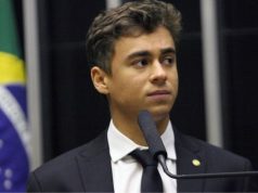 deputado federal Nikolas Ferreira (PL) olhando para o lado, sério, com bandeira do Brasil ao fundo