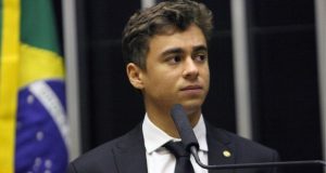 deputado federal Nikolas Ferreira (PL) olhando para o lado, sério, com bandeira do Brasil ao fundo