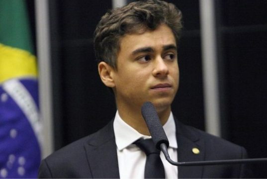 deputado federal Nikolas Ferreira (PL) olhando para o lado, sério, com bandeira do Brasil ao fundo