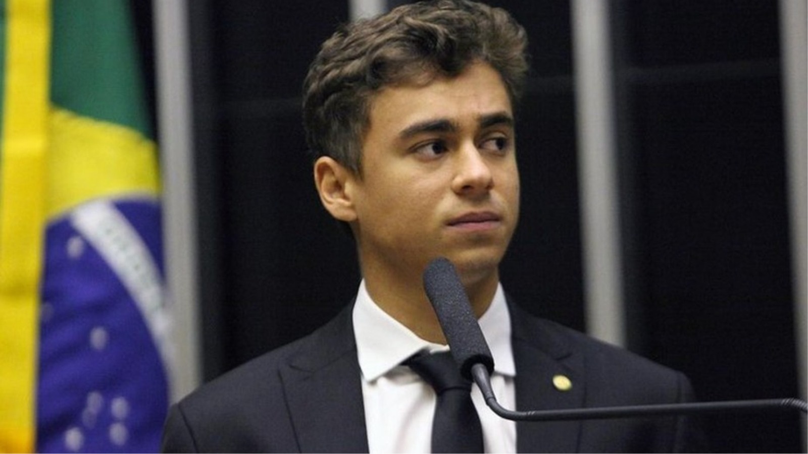 deputado federal Nikolas Ferreira (PL) olhando para o lado, sério, com bandeira do Brasil ao fundo