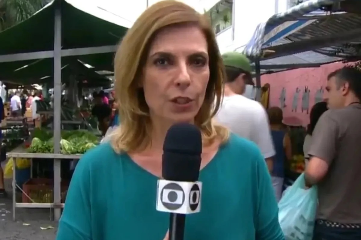 A repórter Zelda Mello de roupa verde, falando em microfone, de cabelo solto, séria, olhando para a câmera