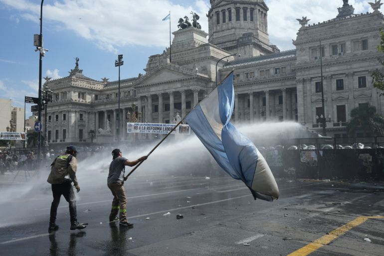 Dois manifestantes, aonde um segura a bandeira da Argentina, enquanto a Polícia, de frente ao parlamento, usa um canhão d'água nos manifestantes.