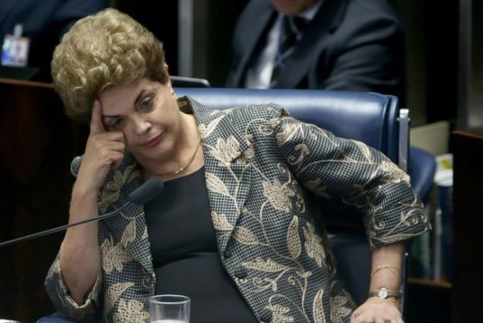 Dilma Rousseff sentada, com o rosto apoiado sobre uma das mãos, séria
