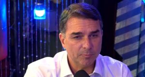 O pré-candidato à Presidência da República Flávio Bolsonaro (PL) falando em microfone, sério, de roupa clara