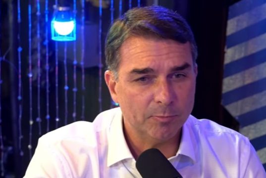 O pré-candidato à Presidência da República Flávio Bolsonaro (PL) falando em microfone, sério, de roupa clara